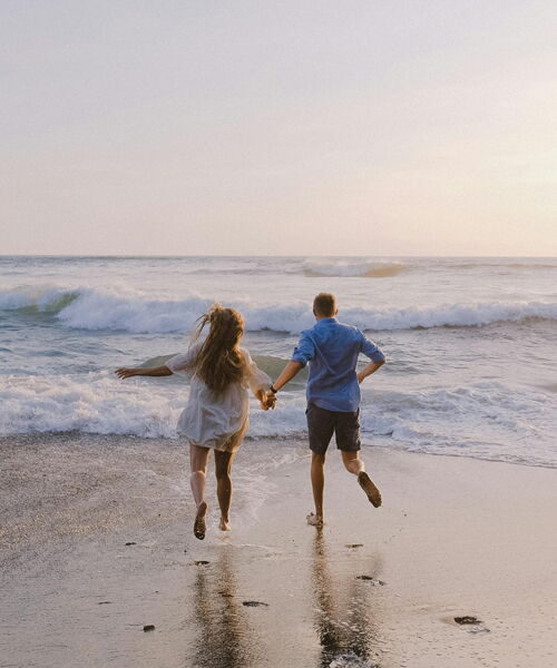 Ein Paar läuft barfuß am Strand auf das Meer mit leichten Wellen zu. Die beiden sind von hinten zu sehen.