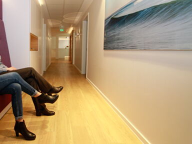 Zwei Personen sitzen im Wartebereich, Blick durch den Flur, nur die Beine sind sichtbar.